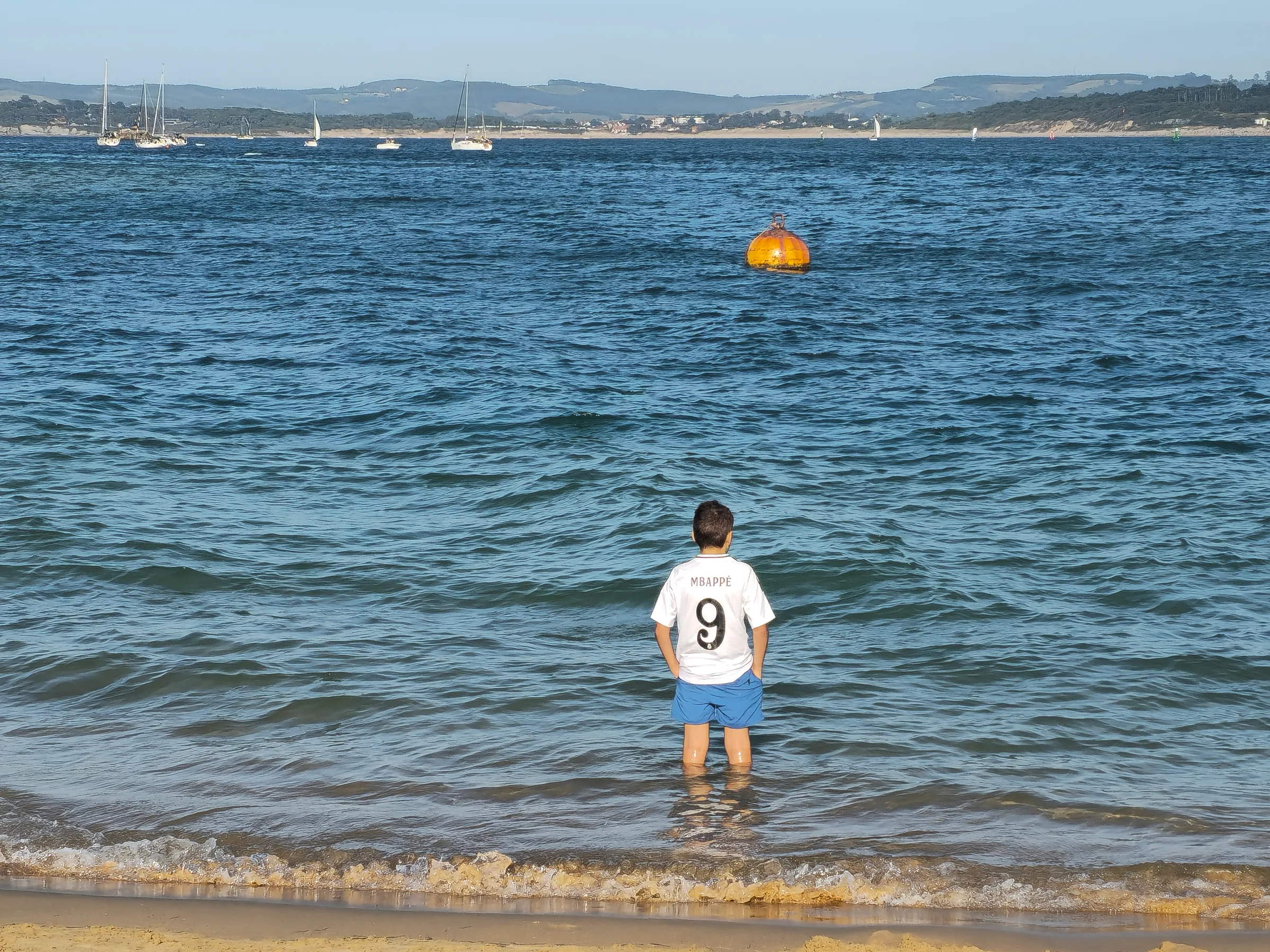 Niño metido en la orilla de la bahía de Santander con veleros al fondo, verano